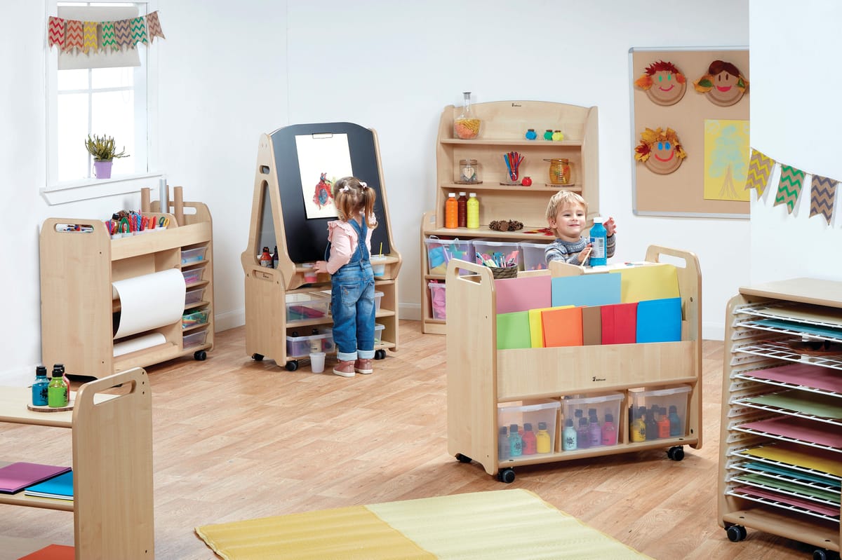 A huge classroom full of different arts and crafts furniture pieces. A little girl is drawing on a chalkboard which has a piece of paper attached to it. A boy is retrieving a bottle of paint from a storage trolley.