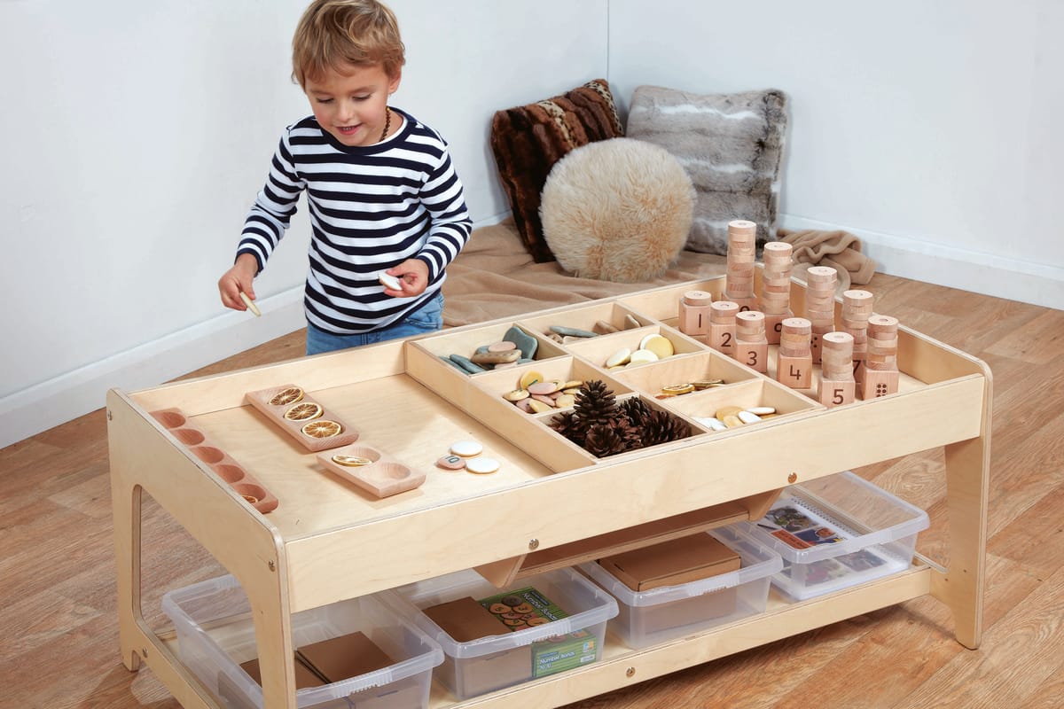 A boy is playing with a variety of props on a wooden table. The table has two levels. The first level contains clear tubs to store objects in and the second level is split into subsections, promoting investigative play.