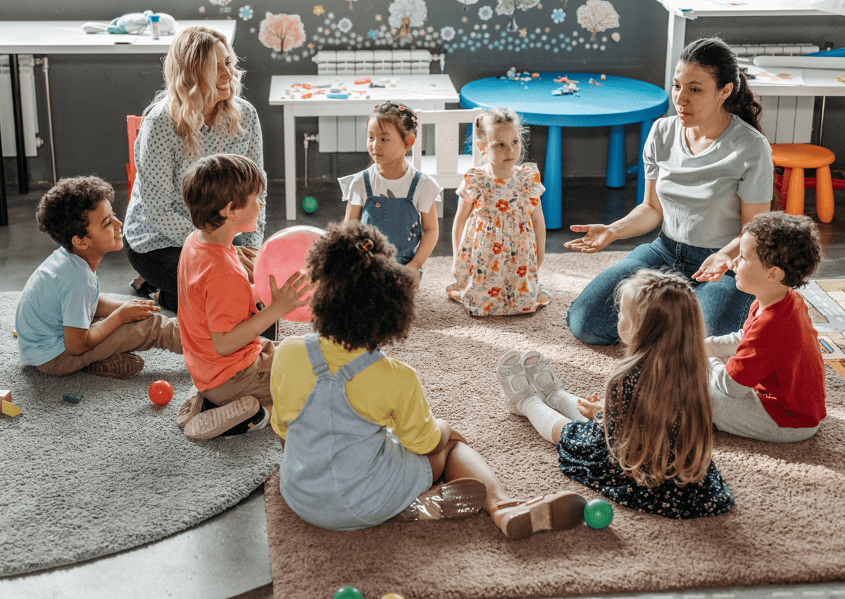 A group of children are sat on the floor with their teacher. The teacher is talking to them as an Ofsted inspector listens into the conversation.