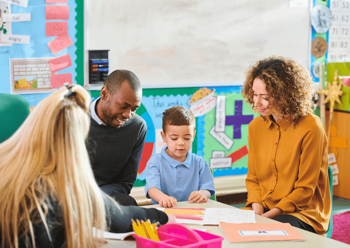 Two Ofsted inspectors are talking to a small child as the child is carrying out an activity. The teacher can be seen too.