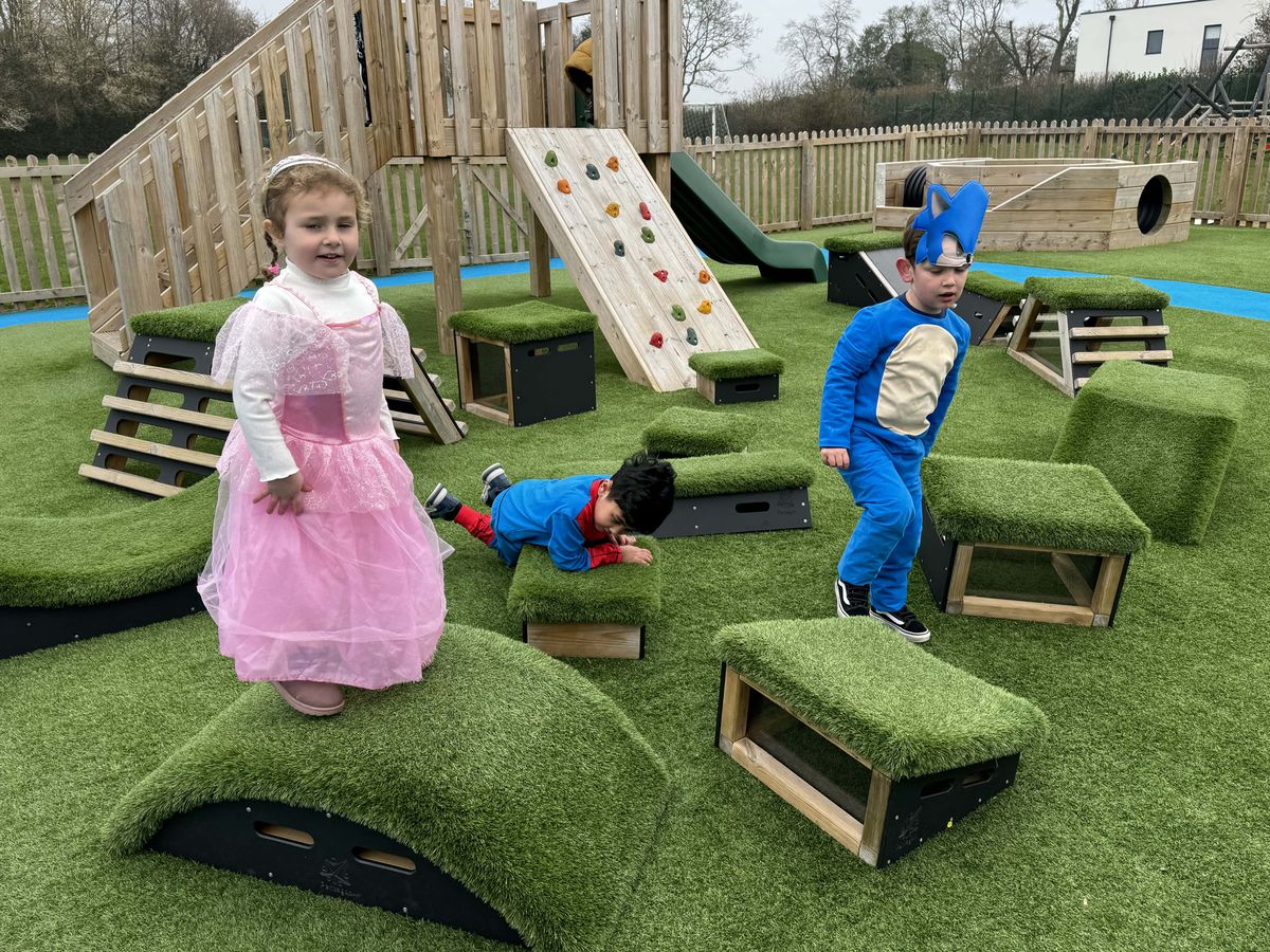 A group of children are playing on a set of Get Set, Go! Blocks, that have been placed on artificial grass, next to a playframe. The children are enjoying themselves and are having fun.