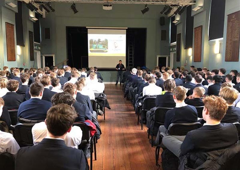 A school assembly hall is full with children as a man is performing a presentation at the front of the hall. The children are all watching the assembly