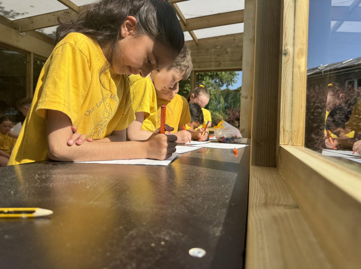 A line of children are sat inside an outdoor classroom and are leaning against a table and writing.