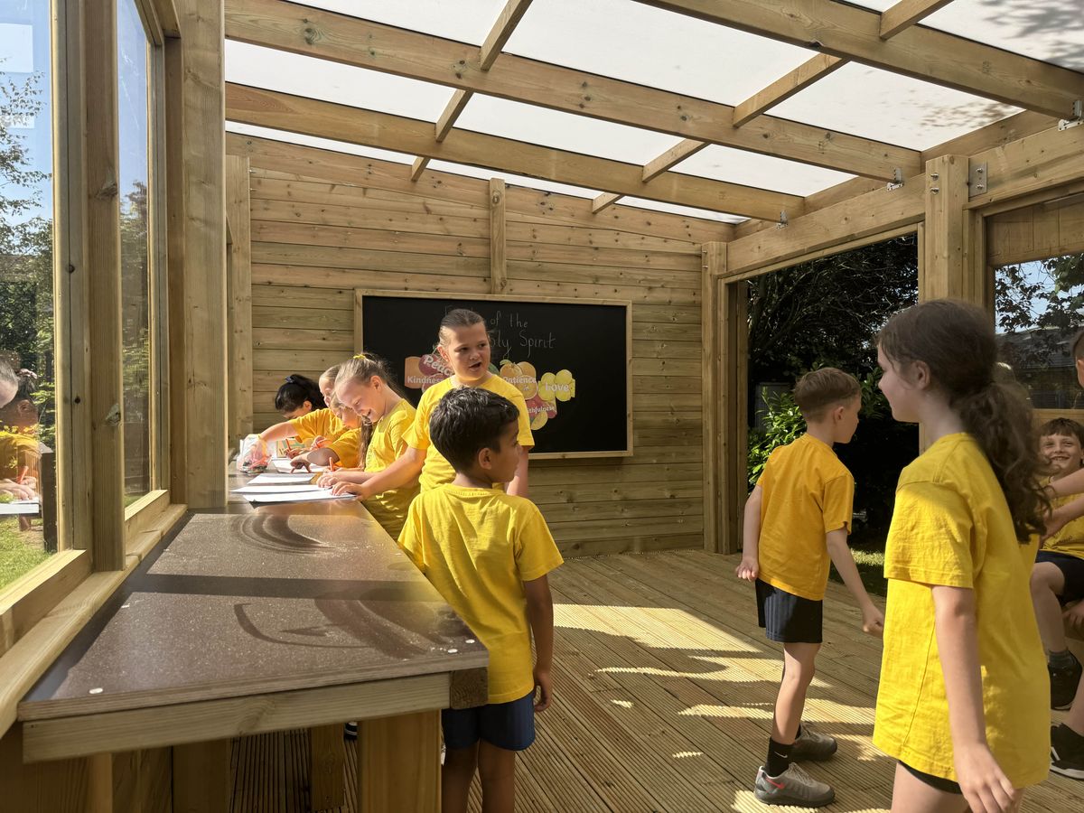 A group of children are stood in an outdoor classroom and are working on a project. The children are walking around and asking each other questions.