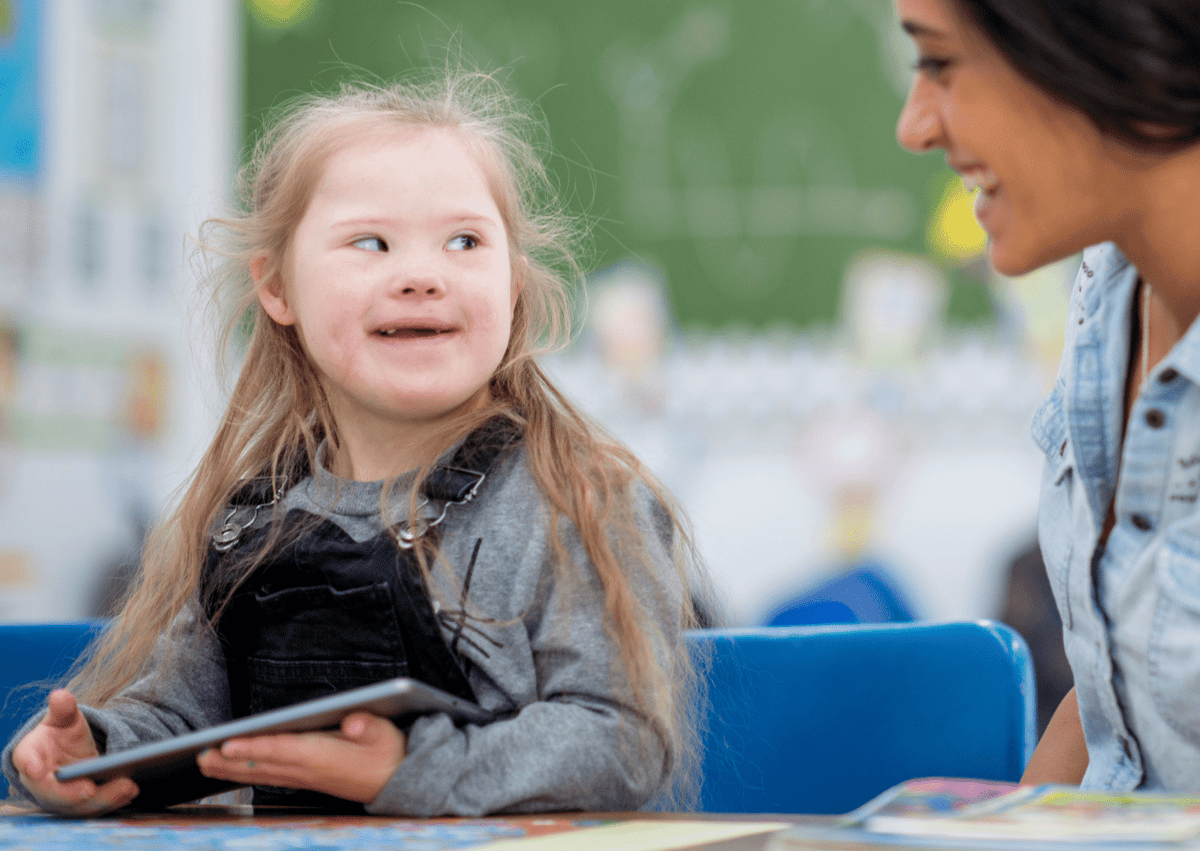 A little girl with SEN is working with a teaching assistant. The girl is smiling and is looking directly at the teaching assistant, who is reciprocating the happiness and joy.