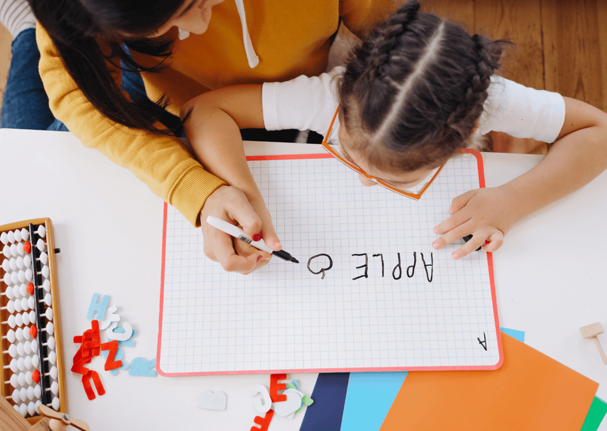A teaching assistant and a child are writing on a whiteboard. They have written the word "Apple" together.