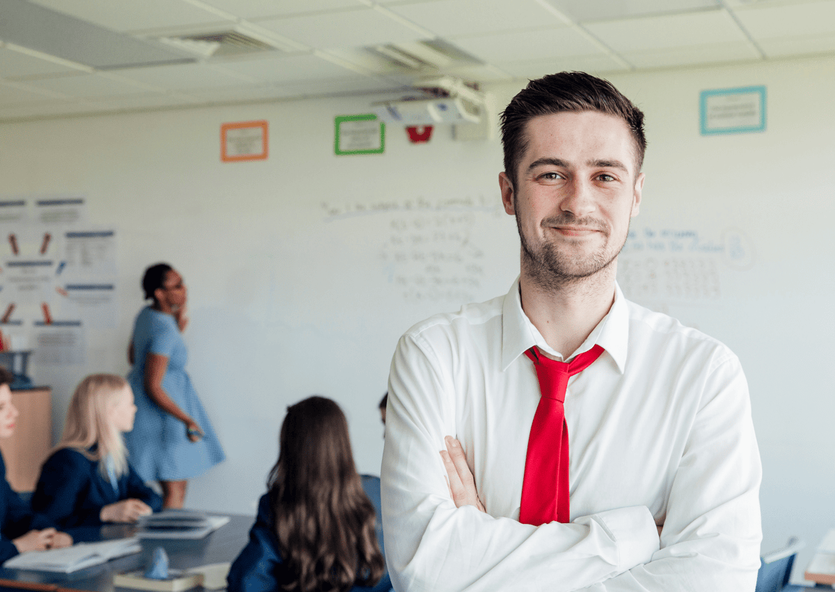 A male teaching assistant, who is wearing a white shirt with a red tie, is looking at the camera and crossing his arms. He has a smile on his face as he looks at the camera, whilst his class are working hard in the background.