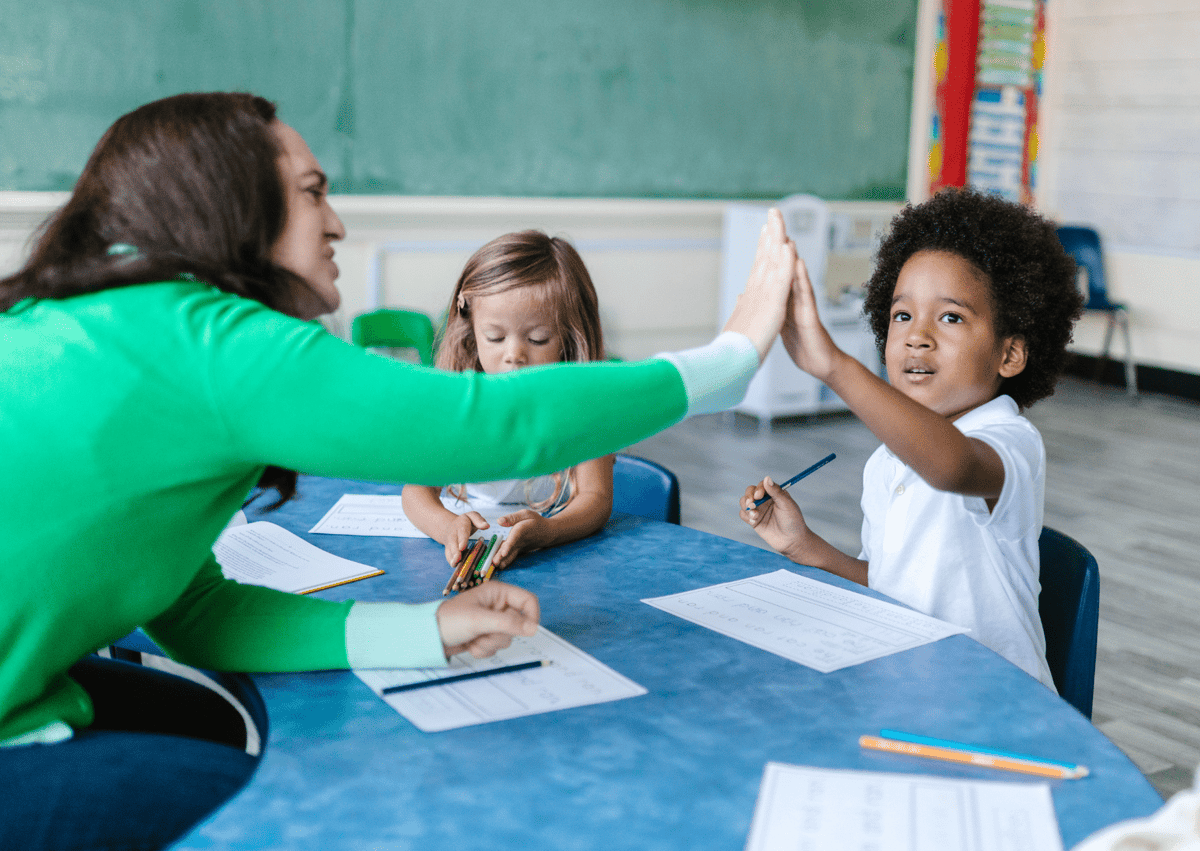 A teaching assistant is sat at a round table with a small group of children. She is leaning over the table and is giving a small boy a high-five.