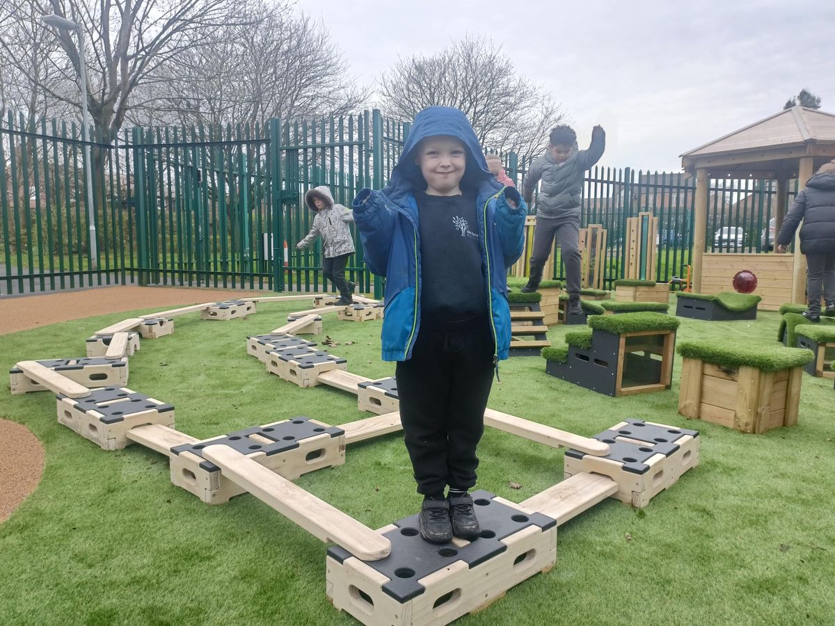 A little boy is stood on top of a Play Builder block as he smiles at the camera.