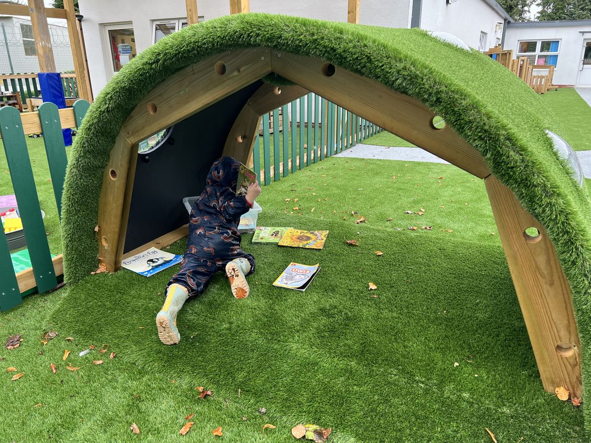 A little girl is lay beneath an artificial grass tunnel and is reading a book.