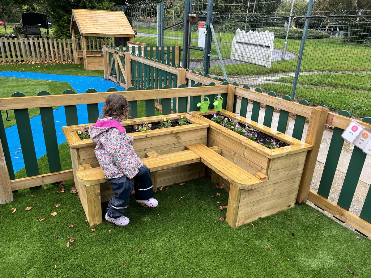 A little girl is sat on a bench and is looking into the planter behind her, watching the flowers grow.