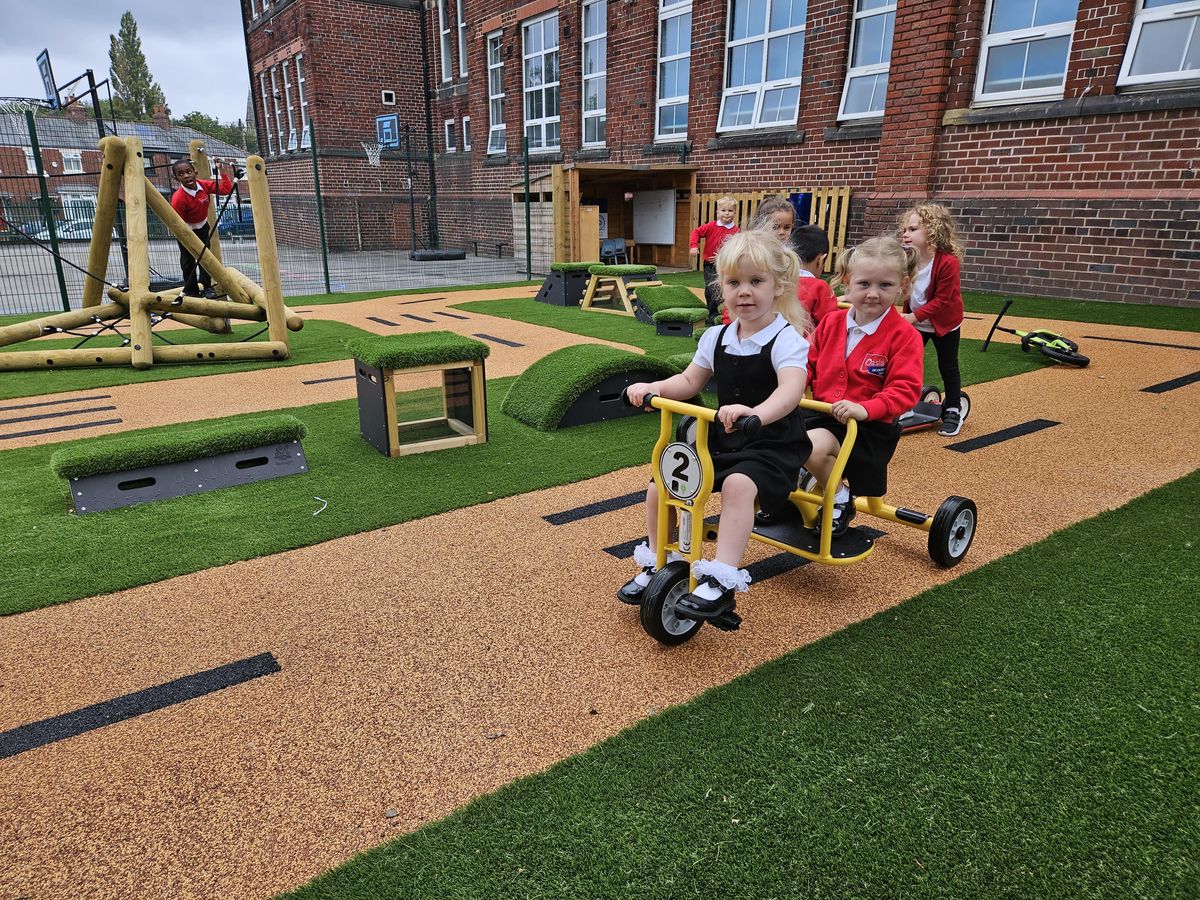 A group of young children are playing on a playground with different pieces of playground equipment on it. Two children are on a tricycle and are looking into the camera and smiling.
