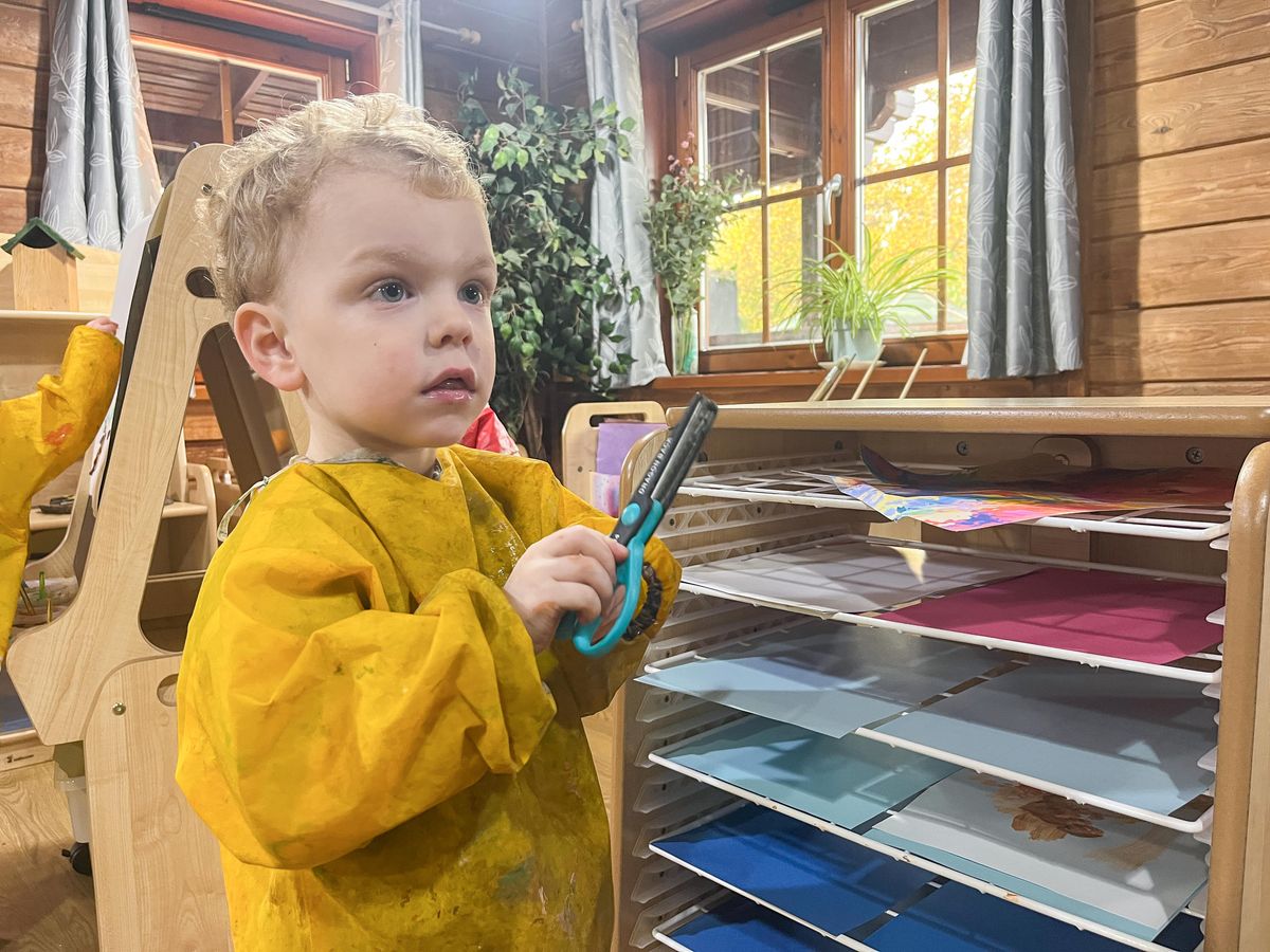 A little boy is holding a pair of safety scissors as he stands next to the A3 drying rack.