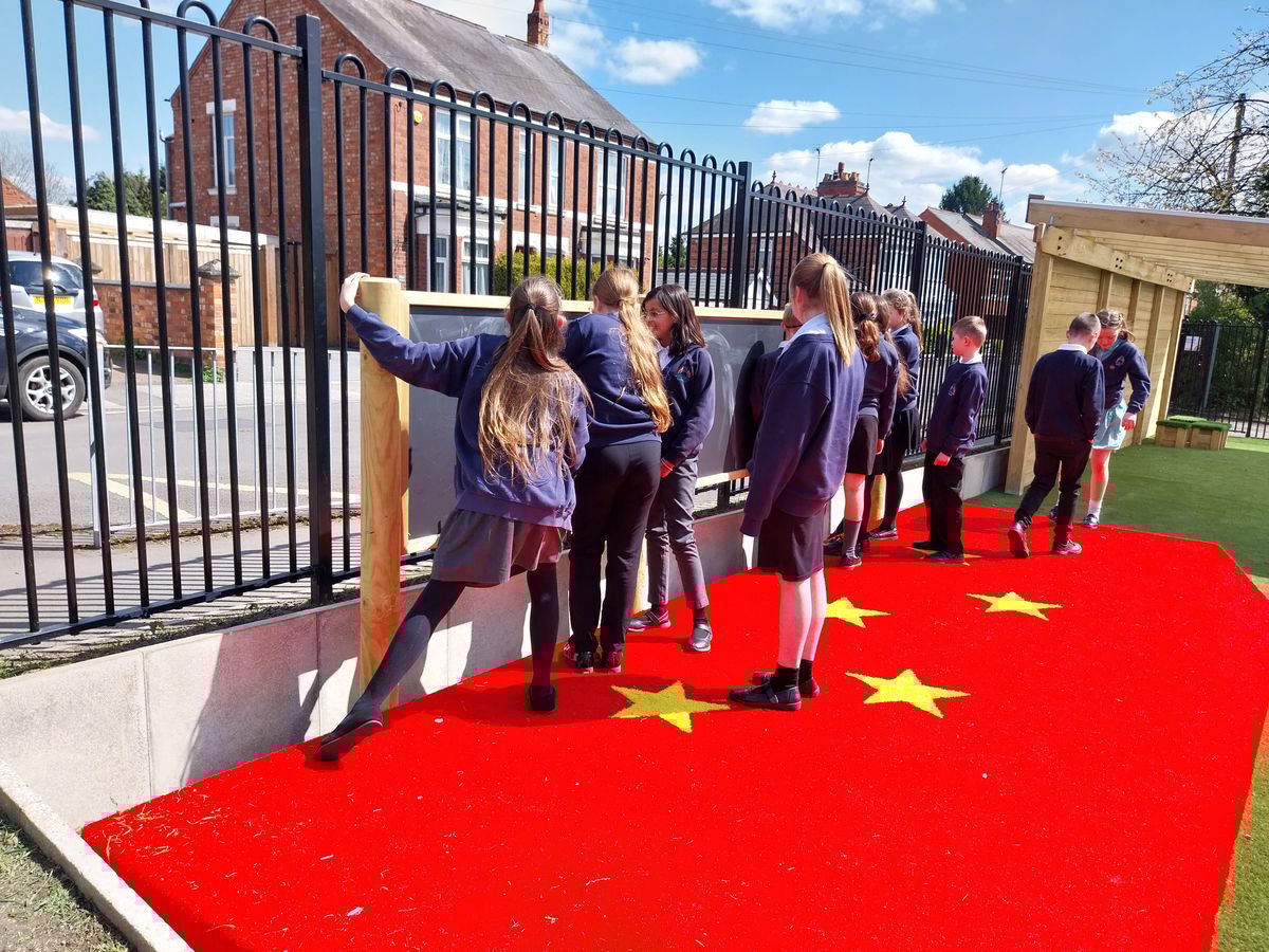 A group of children are stood on an outdoor performance stage as they draw on a chalkboard and prepare their class for a Christmas play.