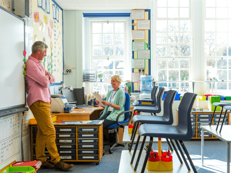 Two teachers are inside a classroom as it's empty, talking about their days.