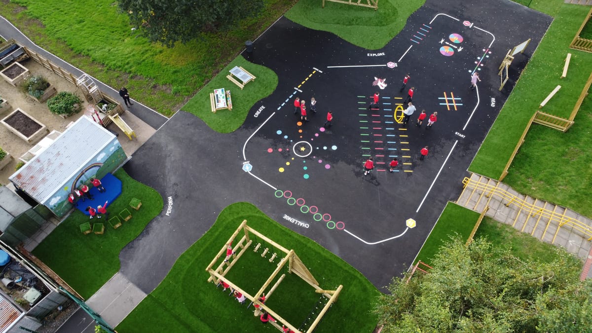 A tarmac playground with thermoplastic playground markings and artificial grass spots present. A variety of playground equipment can be seen around the area.