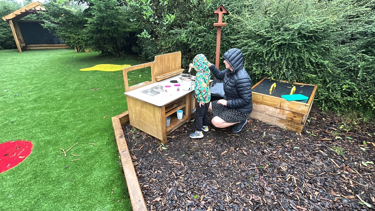 A little boy is playing with a mud kitchen as a teacher squats next to him and helps him.