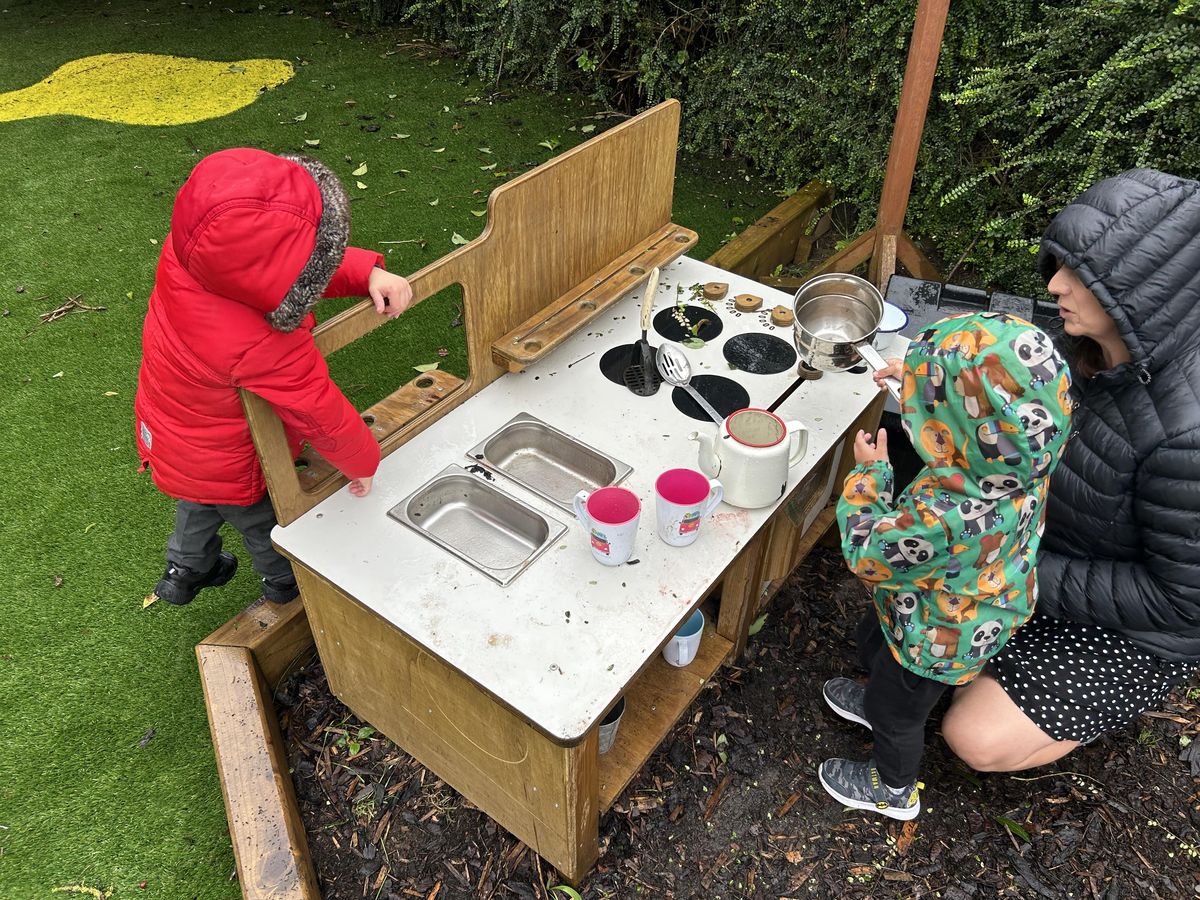 Two children are stood at a mud kitchen and are playing with a variety of pieces of cooking equipment. A teacher is stood beside the children and is guiding them.