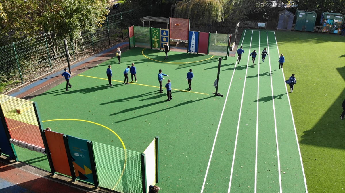 A group of children are playing sports on a MUGA, whilst another group are running on a polymeric running track to the side of the MUGA.