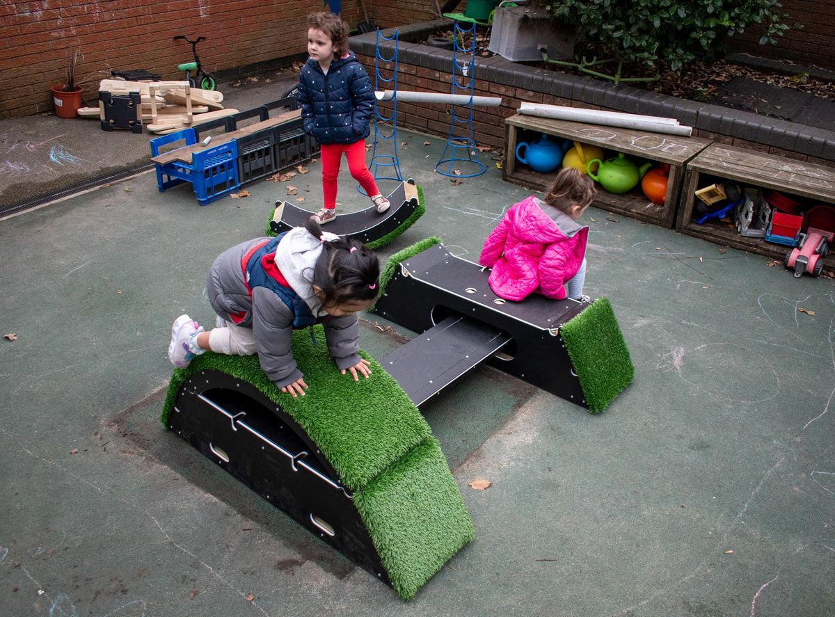 2 children are climbing and playing on the Rockies, a piece of play equipment with a wobble board in-between two ramped blocks.