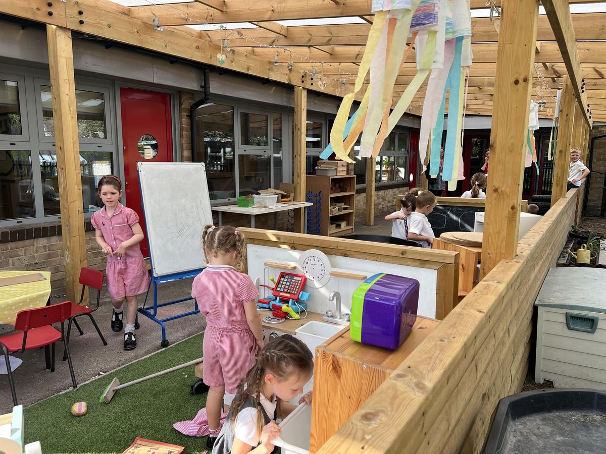 Children stood underneath an outdoor canopy, rooting through a variety of playground storage options to find treasure hunt objects.