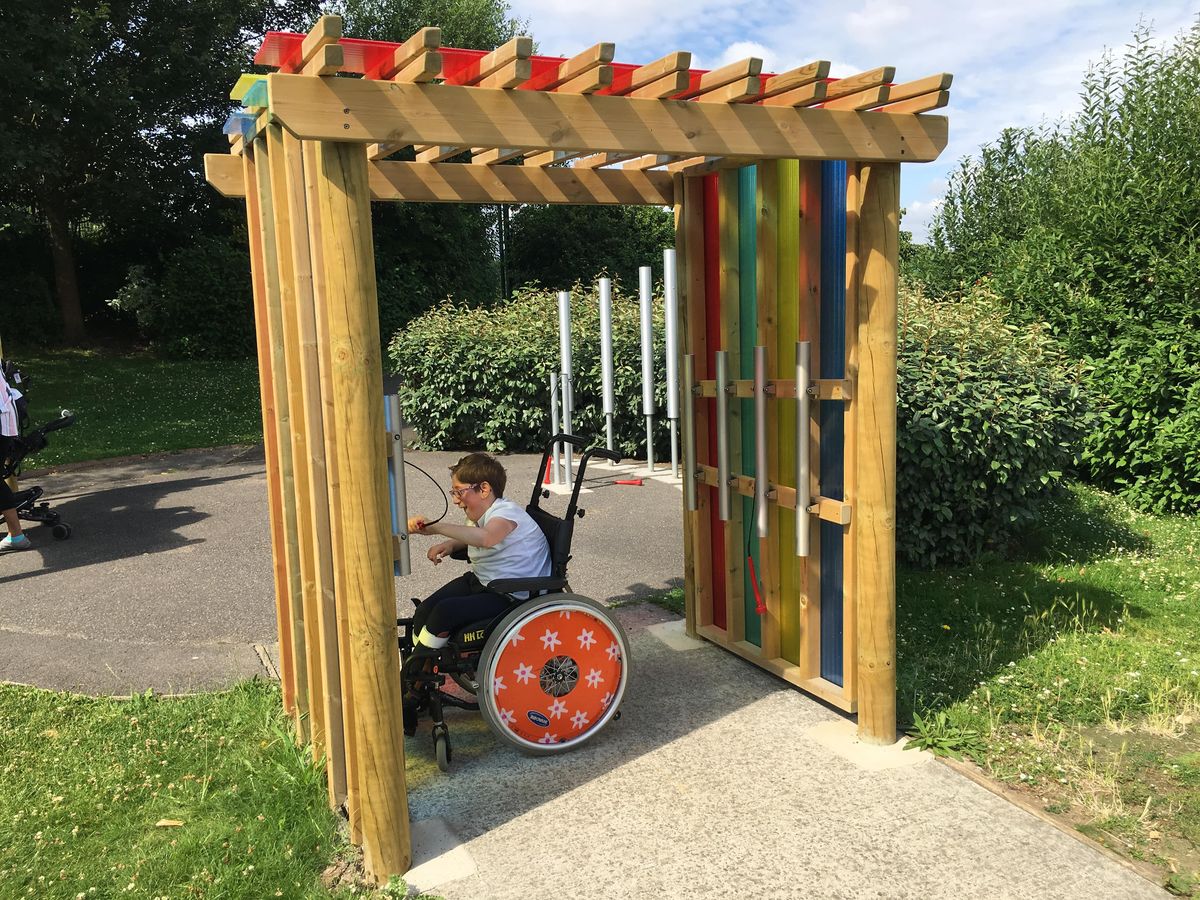 A child in a wheel chair is underneath the sensory arbour and is playing with a wall mounted chime.