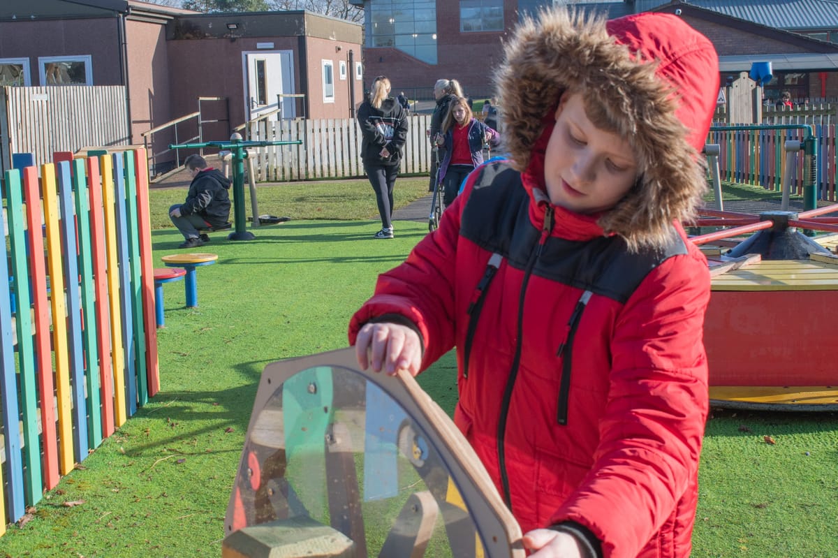 A little boy is playing with a sound sensory spinner as children play behind him.