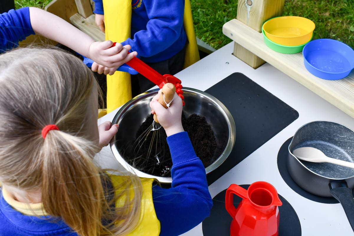 A little girl is mixing a bowl of mud, with the help of her classmates, as she makes a mud pie on a Mud Kitchen.