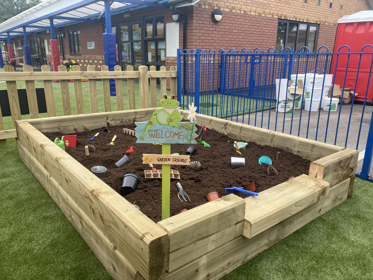 A dig pit that has been filled with mud and a variety of different tools. A sign has been placed at the front, welcoming children.