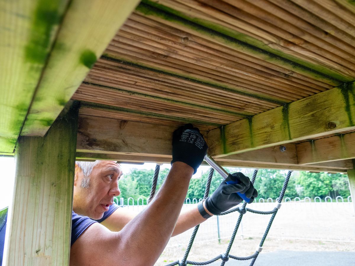 A playground inspector is tightening a bolt on a wooden frame.