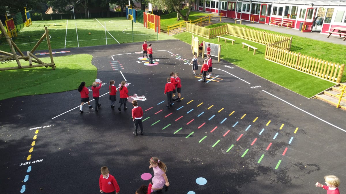 A tarmac playground with a variety of thermoplastic playground markings, ranging in colours. A large group of children are playing with them.