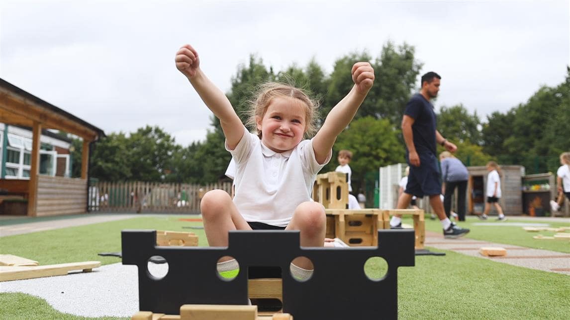 A little girl is sat on a Play Builder plank as she has a lightbulb moment, holding her hands in the air.