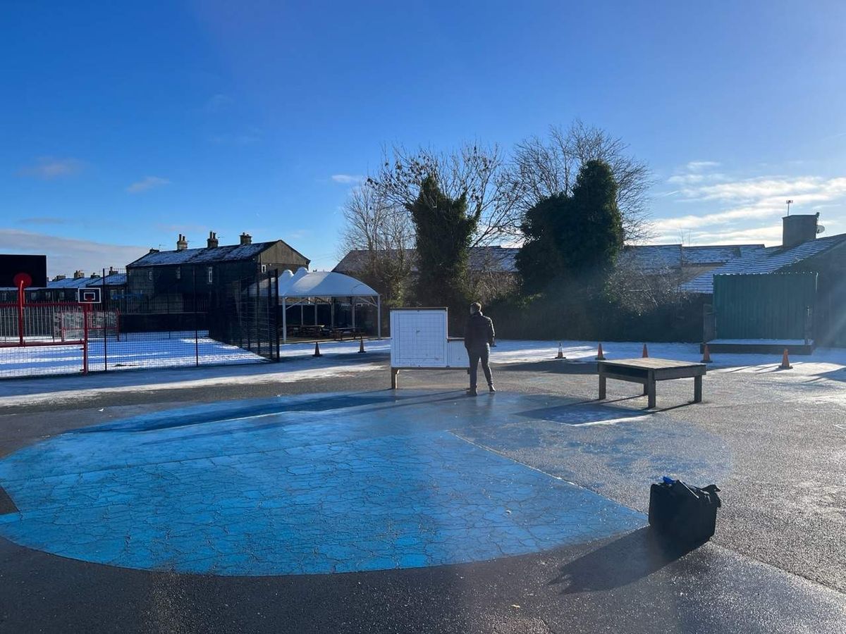 A man is stood on a tarmac playground as he looks around to see the playground is covered in ice and frost, creating a winter image.