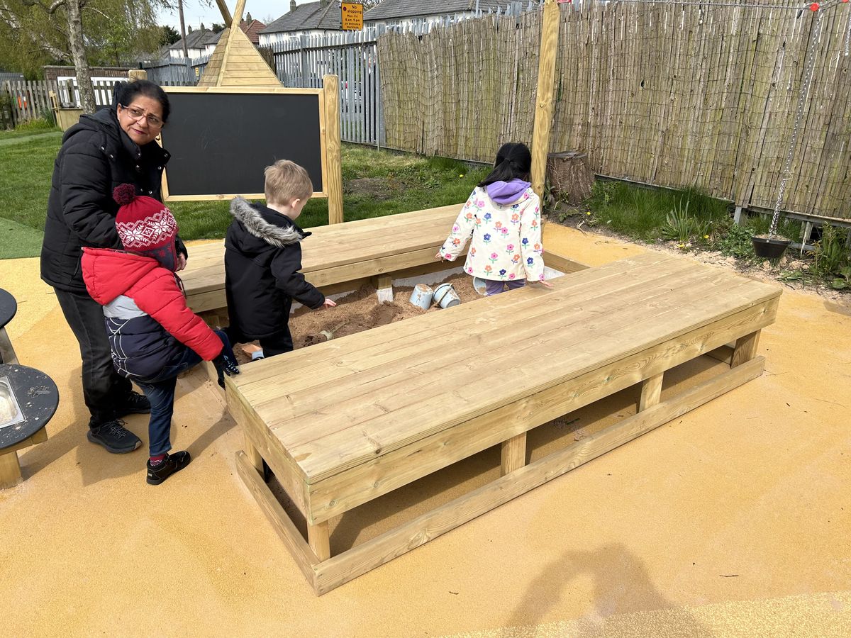 A group of children are being led to a sand pit, as a teacher stands by, watching them.
