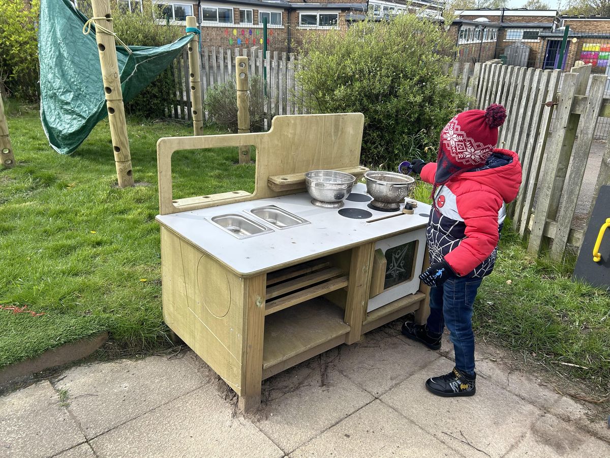 A little boy wearing a coat and a hat is playing with a mud kitchen as he stands in a playground.