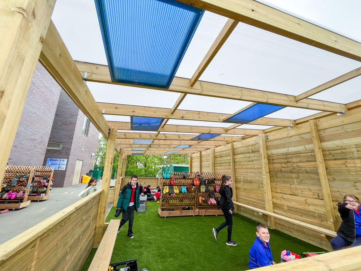 A canopy with coloured roofing and an open space, with loads of children inside of it taking shelter from rain.
