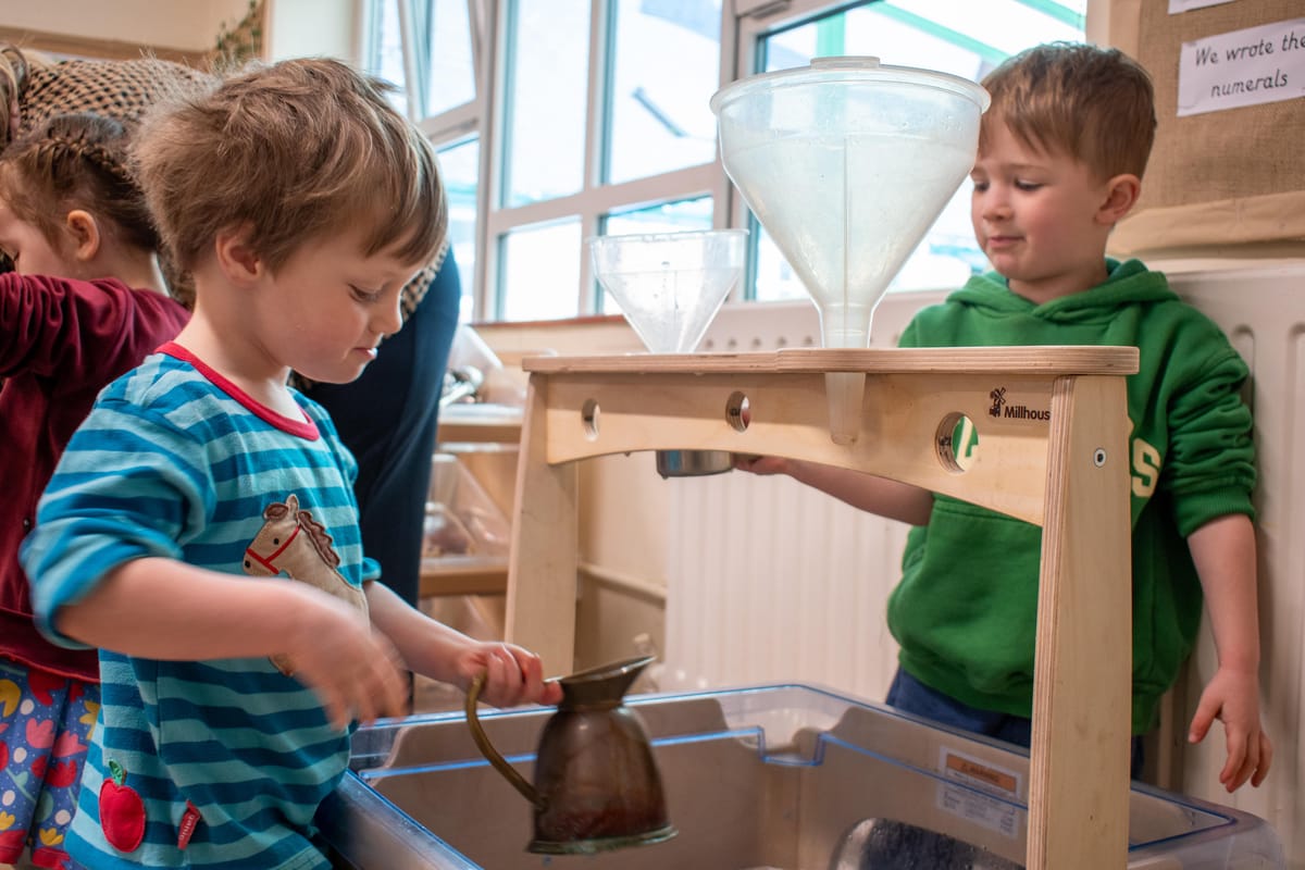 Two little boys are stood next to an indoor water station as they pour water inside the structure.