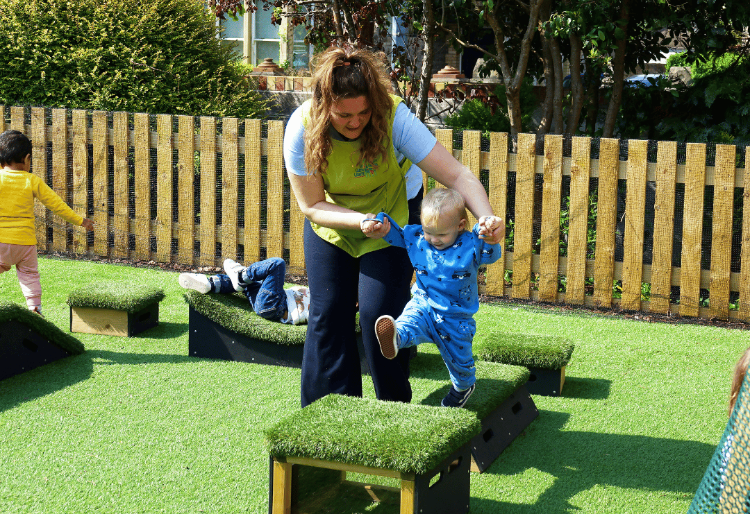 An early years teacher is holding the hands of a child as she helps them navigate an active play course.