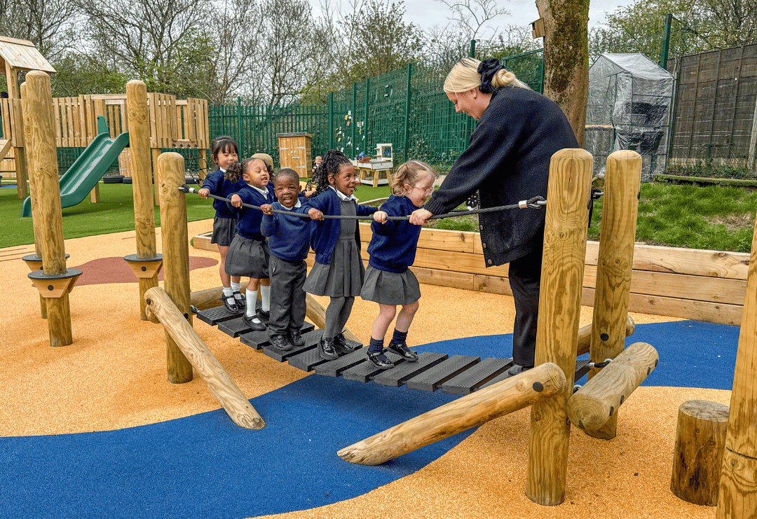 A line of children are stood on a log bridge as their early years teacher is stood on the other side of the bridge.