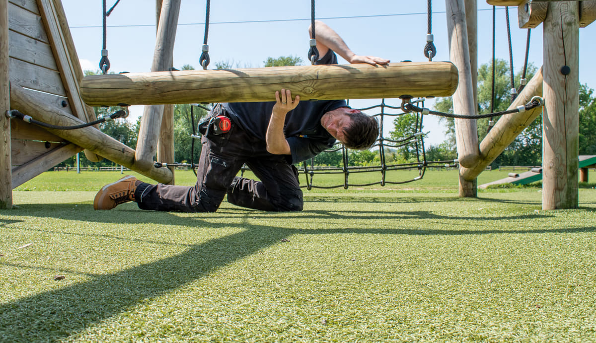 A playground inspector is looking beneath a log swing for any signs of dirt or rot.