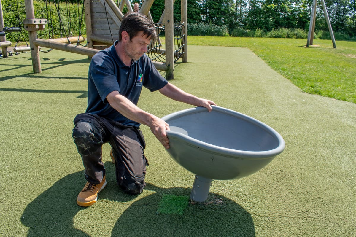 A playground inspector has his hands on a spinning chair and is rocking it, discovering the piece of play equipment has become loose from the ground.