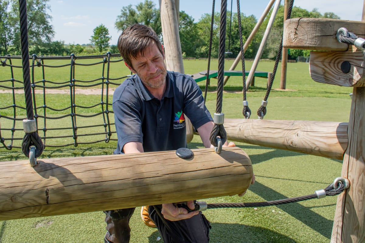 A playground inspector is checking the metal hoops that connect the rope to the log swing for rust.