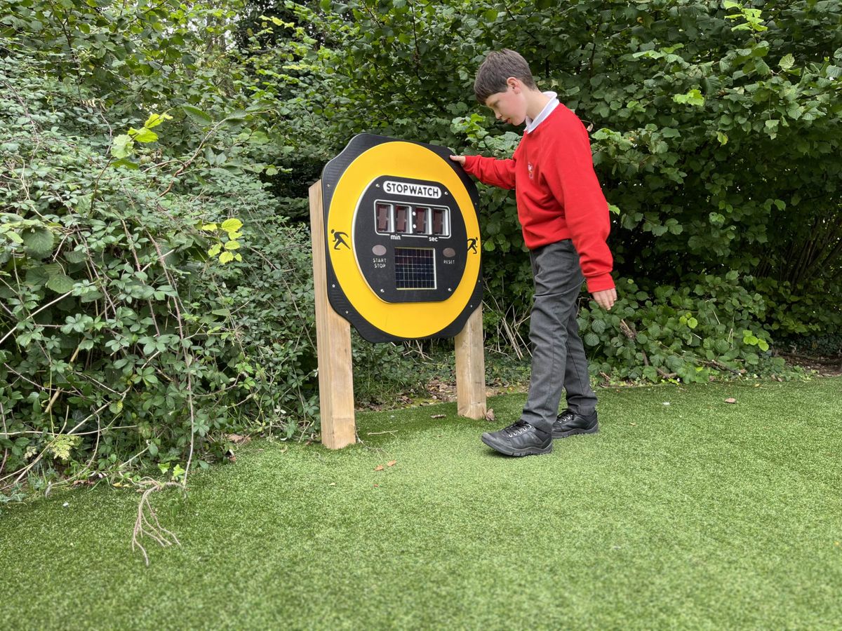 A little boy is stood beside the giant stopwatch on wooden poles as he is looking at the timer.