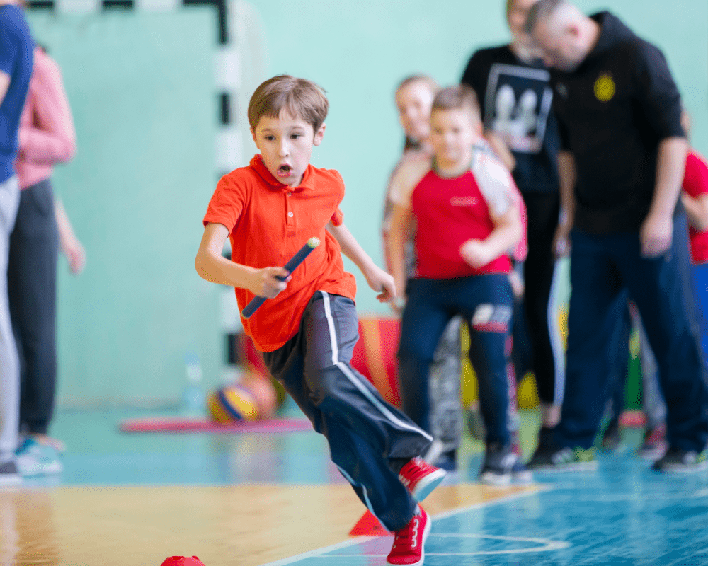 A boy is running around some cones inside a sports hall as he holds a relay baton.