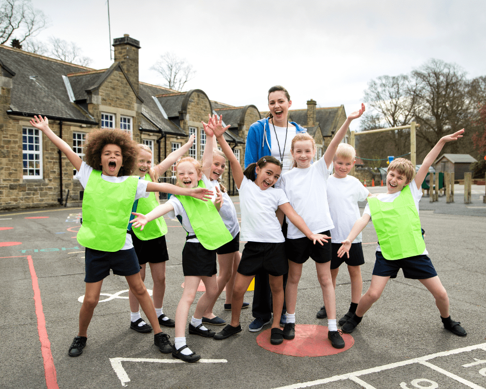 A group of children are stood in front of a teacher and are posing happily towards the camera in their PE kits.