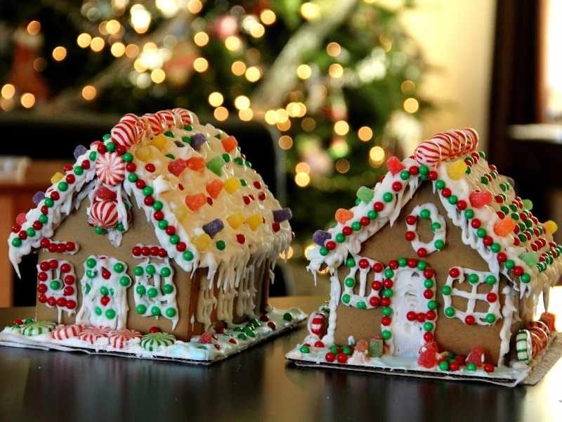 Two gingerbread houses are placed on a table, fully decorated and in front of a Christmas tree with its lights on.