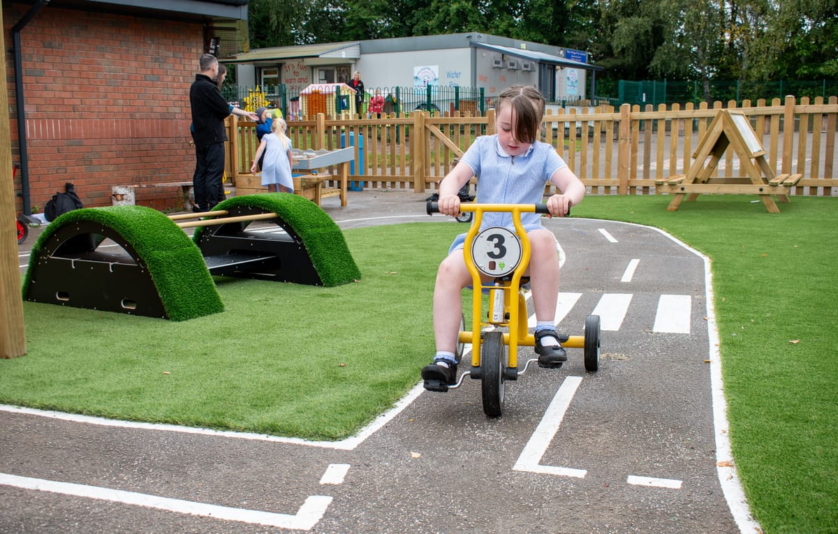 A little girl is on a tricycle and is cycling on a tarmac road graphic with artificial grass surrounding it.