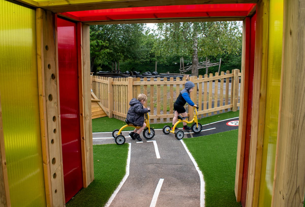 Two children on tricycles are cycling together at different ability levels on a wetpour road.