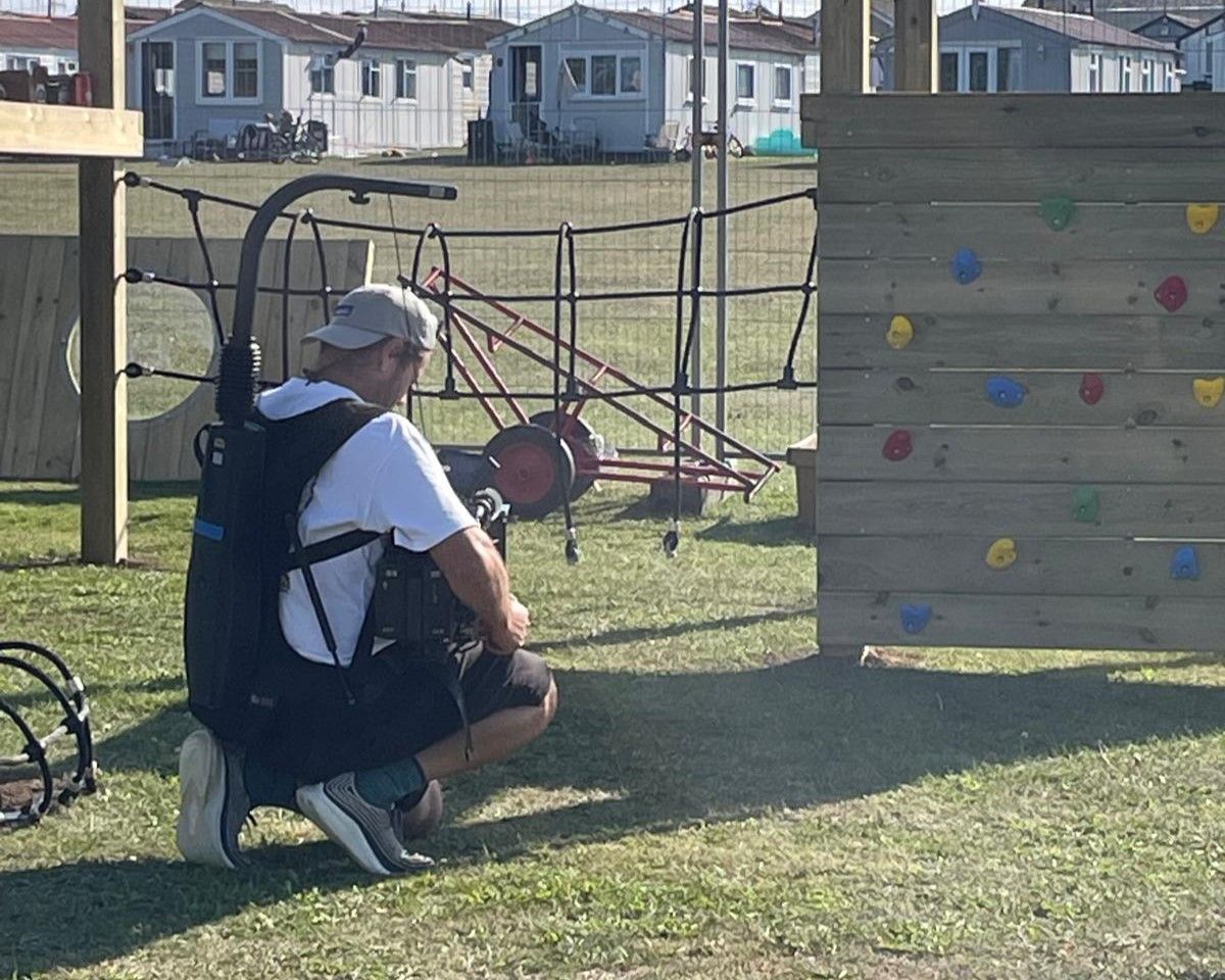 A man is filming a wooden climbing frame whilst kneeling on a natural grass surface.