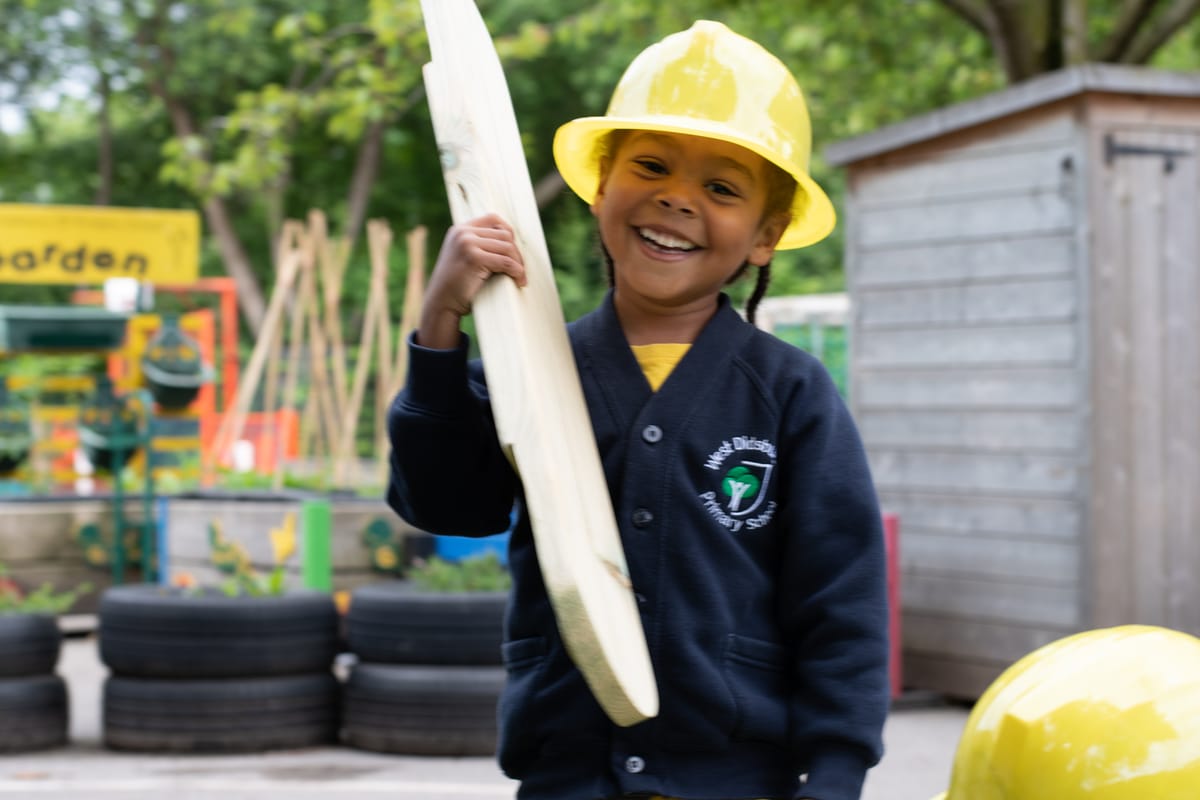 A little girl is holding up a wooden plank from the Play Builder set and is looking at the camera smiling.